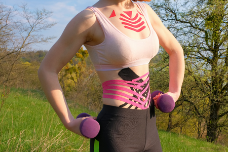 young white girl with kinesio tape on her belly and chest goes in for sports with dumbbells in nature, close up view. Alternative medicine rehabilitationの写真素材