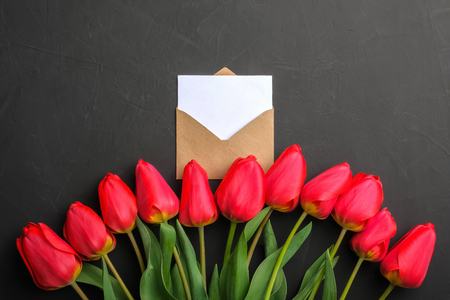 Mockup of fresh red tulips bouquet and white empty greeting card in kraft envelope on dark background with copy spaceの写真素材