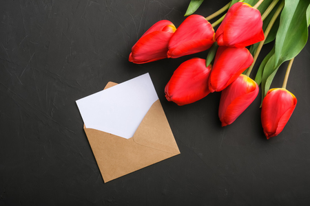 Mockup of fresh red tulips bouquet and blank greeting card in kraft envelope on dark background with copy spaceの写真素材