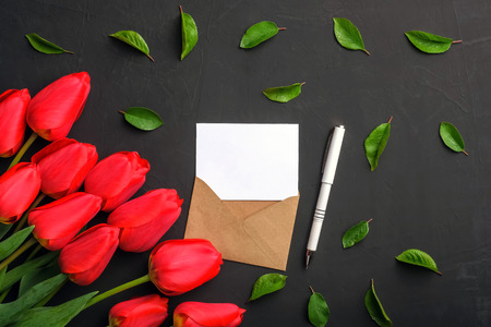 Mockup of fresh red tulips bouquet and greeting card in kraft envelope, pen and scattered leaves on dark background with copy spaceの写真素材