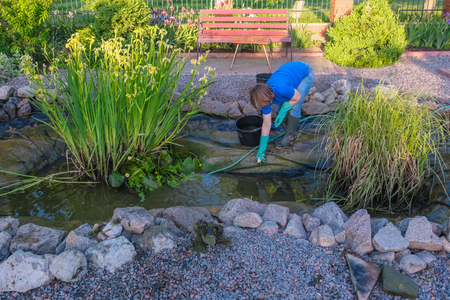 white middle aged  woman cleans fish pond from slime and water plants. Spring and summer seasonal pond care work.の写真素材