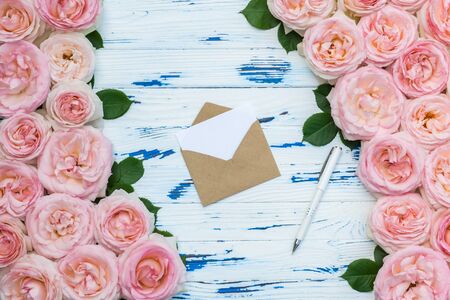 Flowers composition with open craft paper envelope and pen in the frame made of pink roses on aged blue wooden background. Flat lay, top view, copy space.の写真素材