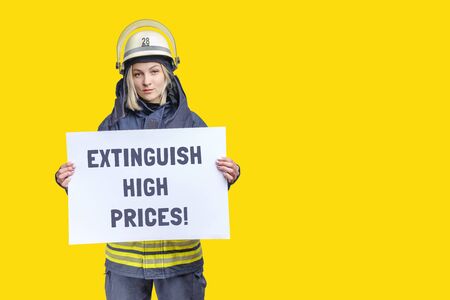 young beauty firefighter wearing uniform and helmet looking to the camera and holding paper sheet with inscription extinguish high prices in hands. isolated on yellow backgroundの写真素材