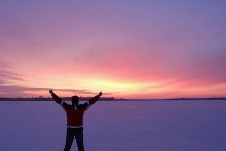 A man with wide outstretched arms in twilight, stands back to the camera on the snowy surface of the frozen lakeの写真素材