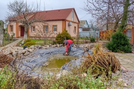 white man cleans a garden pond with a landing net from slime, water plants, falling leaves and catches fish for resettlement in an aquarium. Autumn seasonal pond care before winterの写真素材