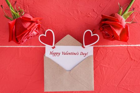 Inscription happy valentine's day on card with craft envelope on red background, roses and hearts on clothespins, closeup viewの写真素材
