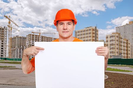 Portrait smiling middle-aged handsome worker wearing orange hard hat holding blank paper sheet in hands, showing finger to copy space for text, on construction site backgroundの写真素材