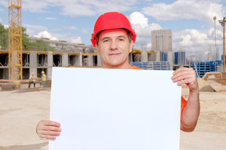Portrait smiling middle-aged handsome worker wearing red hard hat holding blank paper sheet in...
