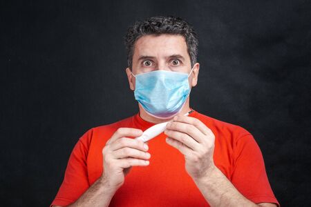 Middle aged caucasian man with medical protective face mask holds thermometer in hands and looking on him with big eyes. MERS Cov middle East respiratory syndrome coronavirus conceptの写真素材