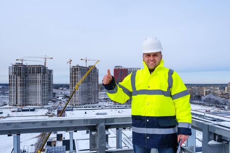 A smiling caucasian builder in a white helmet and a yellow vest gesture thumb up, stands on the roof and inspects construction site.の写真素材
