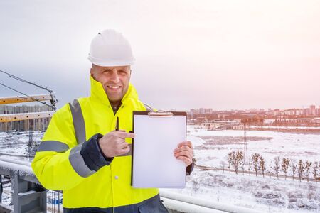 A smiling caucasian engineer wearing white working helmet and a yellow jacket stands on the roof, holds clipboard and pointing finger to empty paper sheet, copy space for your text.の写真素材