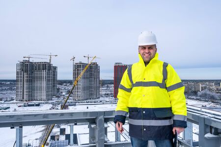 A smiling caucasian engineer wearing white working helmet and a yellow jacket stands on the roof, holds clipboard and looking at the camera in winter.の写真素材