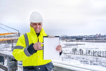 A smiling caucasian foreman wearing white working helmet and a yellow jacket stands on the roof, holds clipboard and pointing finger to empty paper sheet, copy space for your text.の写真素材