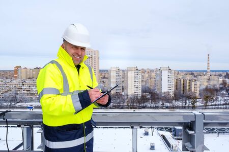 A smiling caucasian foreman in a white helmet and a yellow vest holds clipboard, stands on the roof and make notes on construction site.の写真素材