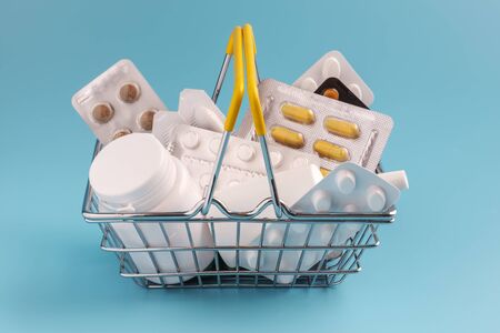 Shopping basket with medicinal pills and tablets closeup. Variety of pills, drugs, spray, syringe, suppositories in mini basket on blue background. Pharmacy Shopping Concept.の写真素材