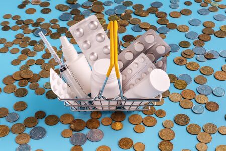 shopping basket filled medicinal tablets,pills and spray bottle on scattered coins on blue background. most expensive drugs or rising drug prices conceptの写真素材