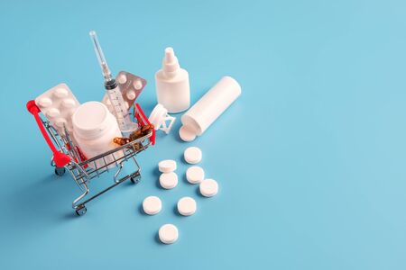 Shopping cart with medicinal pills and scattered tablets. variety of pills, drugs, spay, syringe in mini basket on blue background. Pharmacy Shopping Concept, copy space, view from aboveの写真素材