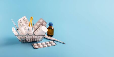 Shopping basket with medicinal pills, spray and syringe. medicine bottle, thermometer and tablets packaging lying near mini-asket on blue background. Pharmacy Shopping Concept, copy space, bannerの写真素材