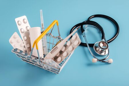 mini shopping basket with medicinal pills, syringe, thermometer and lying near stethoscope on blue background. Pharmacy Shopping Conceptの写真素材