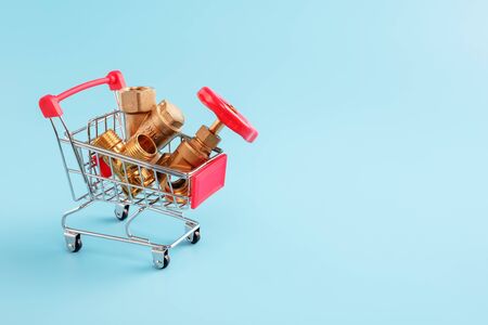 red shopping cart with plumbing fittings closeup. Brass gate valve, y filter and tee in mini basket on blue background. copy space, shopping concept.の写真素材