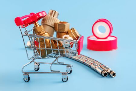 red shopping cart with plumbing parts. Brass gate valve, filter, polypropylene fitting in mini basket. sealing tape and flexible hoses on blue background. pipeline installation, shopping concept.の写真素材