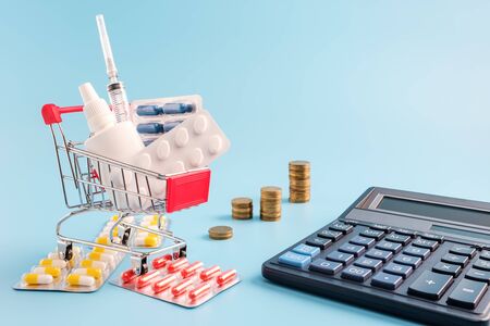 shopping trolley cart filled pills, tablets and syringe, calculator and stacks of coins on blue background, selective focus. most expensive drugs or rising drug prices concept, copy spaceの写真素材