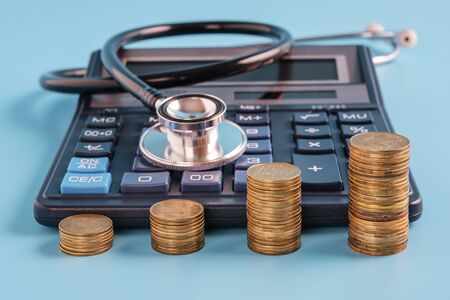 stacks of coins in a row ascending. Calculator and sthetoscope on blue background, selective focus. cost of health care conceptの写真素材