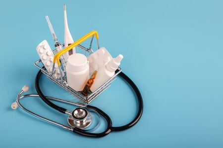 stethoscope lying near mini shopping basket with medicinal pills, syringe and thermometer on blue background. Pharmacy Shopping Concept, copy spaceの写真素材