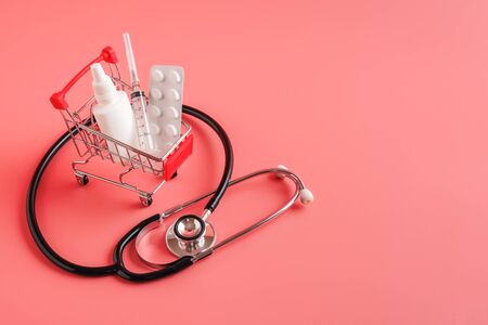 Shopping cart with medicinal pills, syringe and bottle. stethoscope lying near mini basket on pink background. Pharmacy Shopping Conceptの写真素材