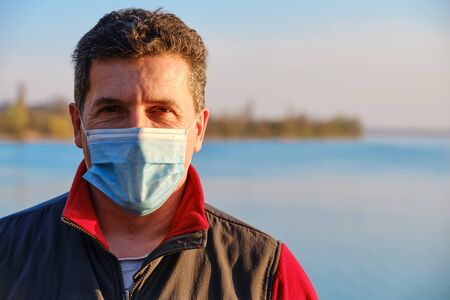 Mature caucasian man wearing protective surgical face mask to prevent coronavirus, on the lake background. Close Up portrait, copy spaceの写真素材