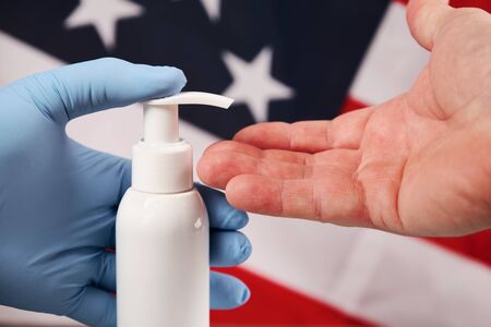 Doctors hand in blue medical glove applying antiseptic alcohol gel on hand without a glove to prevent infections coronaviruses, bacteria and germs, on american flag background. Closeup viewの写真素材