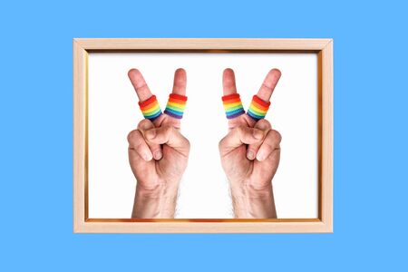 wooden frame with two male hands making the V sign on blue background. fingers wears wristbands painted as rainbow flag. Pride day concept, flat layの写真素材