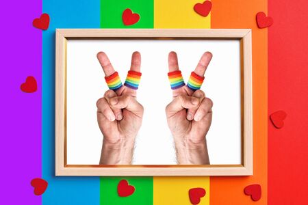 wooden frame with two male hands making the V sign on multicolored rainbow flag background and little red hearts. fingers wears wristbands painted as rainbow flag. Pride day concept, flat layの写真素材