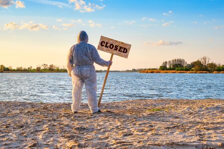 Male wearing protective suit stands with warning sign in hands with inscription closed along the sand beach of the river. Contaminated water, quarantine, virus outbreak, crime scene conceptの写真素材