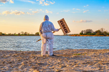 Man wearing protective suit stands with warning sign in hands with inscription closed along the sand...