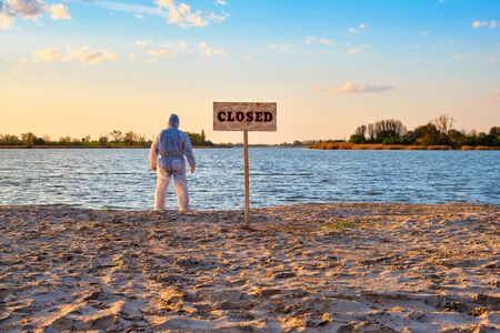 Warning sign with inscription closed along the sand beach of the river and male wearing protective suit stands with his back.の写真素材