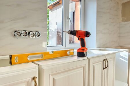 Cordless screwdriver and yellow building level on white kitchen furniture. Light interior, preparing to custom installation processの写真素材