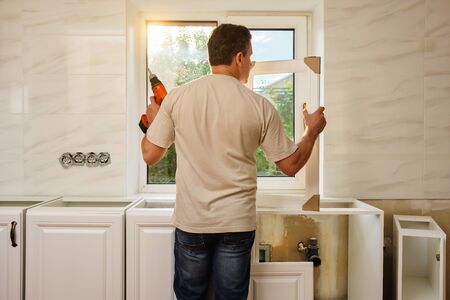 Middle-aged caucasian man stands with his back and holds the kitchen cabinet door and cordless screwdriver, preparing for the process of individual installation in his own house. Quarantine activityの写真素材