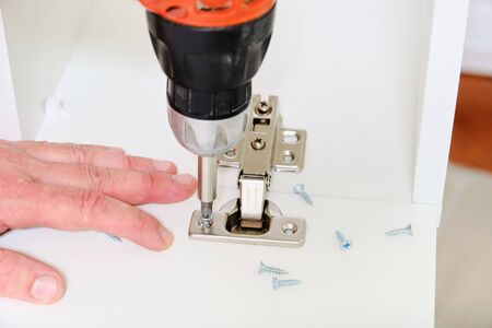 Male hands screwed the furniture hinges into kitchen cabinet with cordless screwdriver closeup.の写真素材