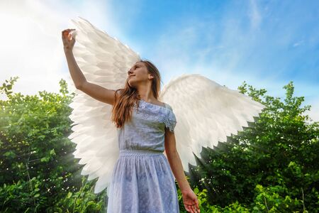 cute caucasian girl with angel wings and in a white dress posing on a background of blue sky.の写真素材