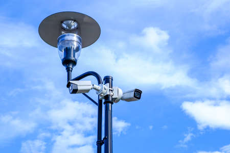 Two white surveillance cameras on the metal lamp post on blue sky background.の写真素材