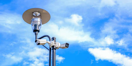 Two white surveillance cameras on the metal street light lamp post outdoor on blue sky backgroundの写真素材