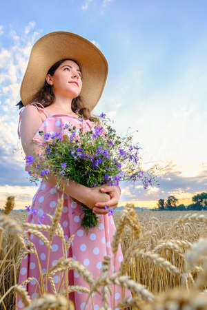 Pretty young girl with long hair holds a wild flower bouquet in wheat field at sunrise.の写真素材