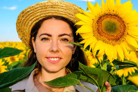 close up portrait of a smiling woman in a straw hat with a sunflower flower in a fieldの写真素材