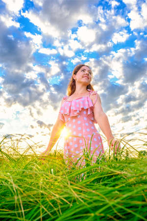 Young smiling caucasian girl standing with outstretched arms in grass on meadow against cloudy sky backgroundの写真素材