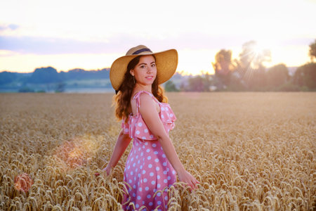 Young woman in straw hat stands turning around on a background of wheat field and smiling