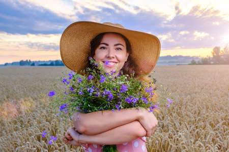 Pretty young woman in straw hat holds a wild flower bouquet in wheat field at sunriseの写真素材