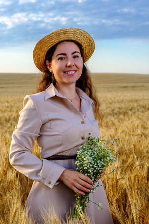 Attractive young long-haired woman in straw hat smiling and holds a wild flower bouquet in wheat field at sunriseの写真素材