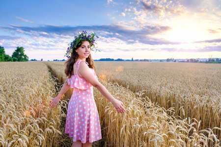 Young woman with flower wreath on head stands turning around on a background of wheat field and smilingの写真素材