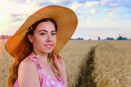 Portrait of smiling young long-haired Ukrainian woman in a straw hat in a wheat field at dawnの写真素材
